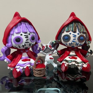 Puede incluir: Dos muñecas con capucha roja con caras cosidas y cabello morado y verde. Las muñecas sostienen una figurita de lobo blanco. Las muñecas están sentadas sobre una superficie negra.