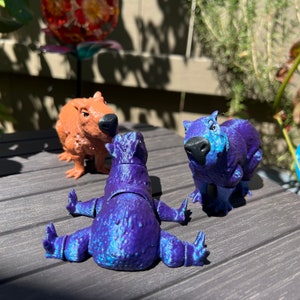 May include: Three capybara figurines, one brown, one purple, and one blue, are posed on a wooden surface. The figurines are detailed and have realistic features.