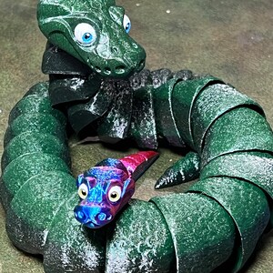 Puede incluir: Una serpiente verde segmentada con grandes ojos azules est&aacute; enroscada en un c&iacute;rculo. Una serpiente segmentada m&aacute;s peque&ntilde;a, azul y rosa con ojos amarillos est&aacute; dentro del c&iacute;rculo.