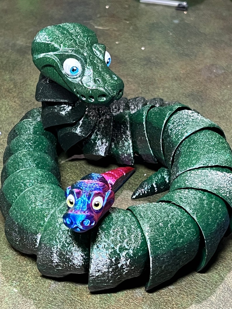 Puede incluir: Una serpiente verde segmentada con grandes ojos azules est&aacute; enroscada en un c&iacute;rculo. Una serpiente segmentada m&aacute;s peque&ntilde;a, azul y rosa con ojos amarillos est&aacute; dentro del c&iacute;rculo.