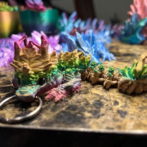 May include: A multi-colored, iridescent, 3D printed dragon keychain with a silver ring. The dragon is detailed with spikes and scales.