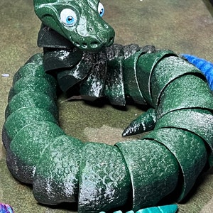 Puede incluir: Un juguete de serpiente verde segmentado con grandes ojos azules. La serpiente est&aacute; enroscada en un c&iacute;rculo con la cabeza mirando al espectador.