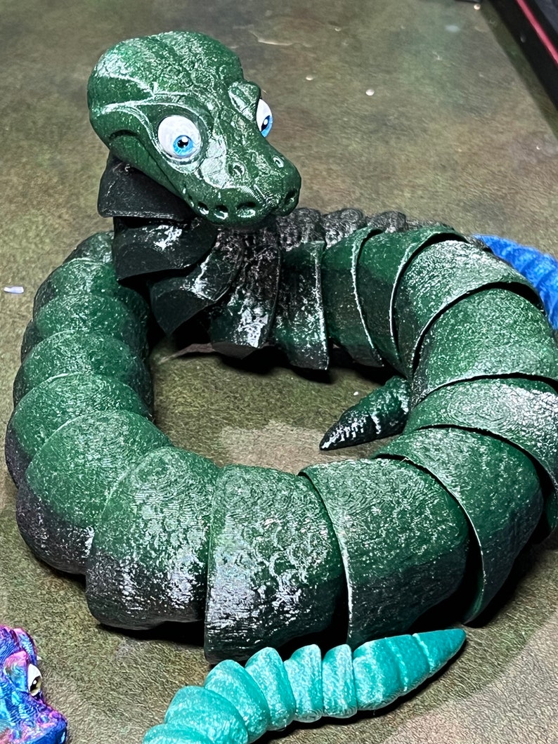 Puede incluir: Un juguete de serpiente verde segmentado con grandes ojos azules. La serpiente est&aacute; enroscada en un c&iacute;rculo con la cabeza mirando al espectador.