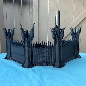 Puede incluir: Un modelo en 3D impreso en negro de una puerta con torres y una puerta grande y ornamentada. La puerta está hecha de muchas piezas pequeñas y afiladas y tiene un aspecto oscuro y amenazante.