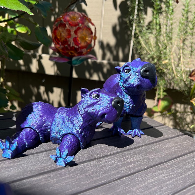 May include: Two purple capybara figurines with iridescent blue highlights. The capybaras are posed in a playful manner, one lying down and the other standing.