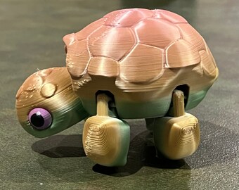 Puede incluir: Una tortuga de juguete impresa en 3D con un esquema de color rosa, verde y dorado. La tortuga tiene un caparazón y patas detalladas.