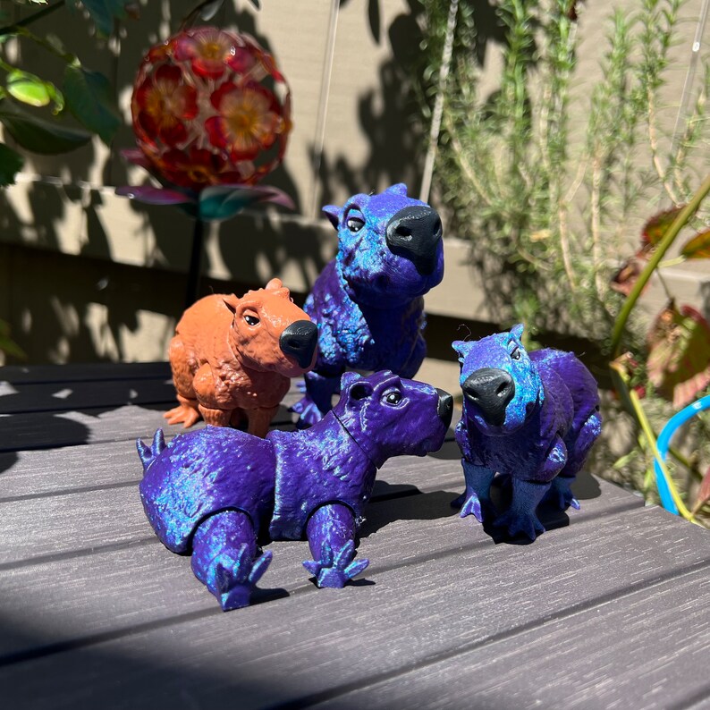 May include: Four small figurines of capybaras, one brown and three purple. The purple capybaras have a shimmery finish.