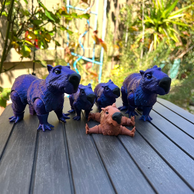 May include: Five small capybara figurines, four are purple and one is brown. The figurines are arranged on a wooden surface.