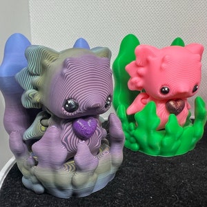 Könnte beinhalten: Zwei 3D-gedruckte Figuren, eine lila und eine rosa, mit einem herzförmigen Design. Die lila Figur steht auf einem Sockel mit einem wirbelnden Muster, während die rosa Figur auf einem grünen Sockel mit einem Blattdesign steht.