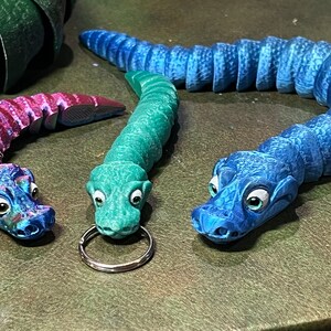 Puede incluir: Tres llaveros de juguete de cocodrilo de colores. Uno es azul, otro es verde y otro es morado y rosa.