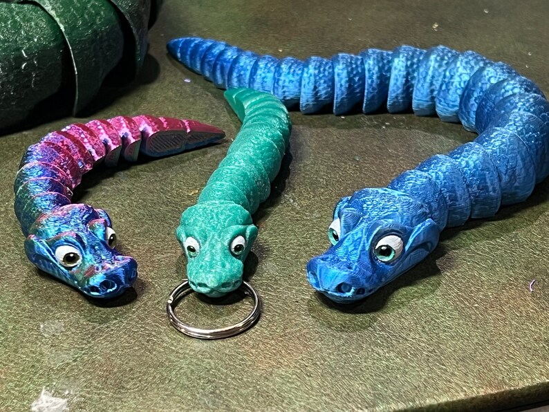 Puede incluir: Tres llaveros de juguete de cocodrilo de colores. Uno es azul, otro es verde y otro es morado y rosa.