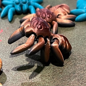 May include: Close-up of several 3D-printed spider figurines. The spiders are primarily brown with black accents on their legs and bodies. Some spiders are teal. The figurines have detailed textures and are arranged on a surface.