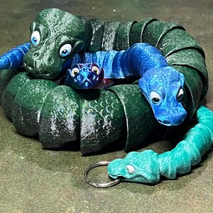 May include: A set of three green, blue, and teal snake figurines. The snakes are curled up together, with the largest snake in the center. The snakes have large eyes and are made of a textured material.
