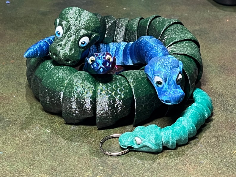 Puede incluir: Un conjunto de tres figuras de serpientes verdes, azules y turquesa. Las serpientes est&aacute;n enroscadas unas alrededor de otras, con la serpiente m&aacute;s grande en el centro. Las serpientes tienen ojos grandes y est&aacute;n hechas de un material texturizado.
