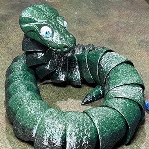 Puede incluir: Un juguete verde, segmentado, con forma de serpiente con ojos grandes y azules. El juguete est&aacute; hecho de un material texturizado similar al caucho y est&aacute; enrollado en un c&iacute;rculo.