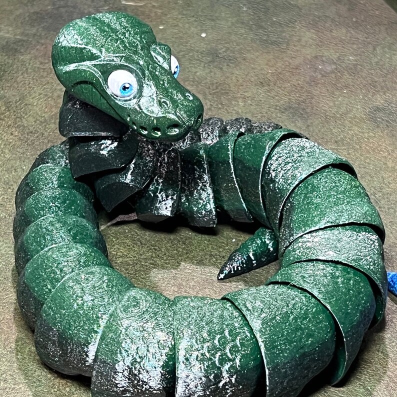 Puede incluir: Un juguete verde, segmentado, con forma de serpiente con ojos grandes y azules. El juguete est&aacute; hecho de un material texturizado similar al caucho y est&aacute; enrollado en un c&iacute;rculo.