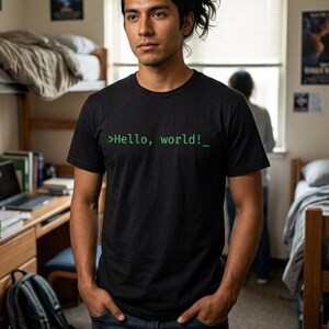 May include: Black t-shirt with the text ">Hello, world!_" in green. The shirt is worn by a person standing in a room with a bunk bed and desk. The person is wearing blue jeans.