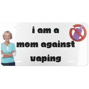 Op de afbeelding: Een wit kenteken met zwarte tekst die "i am a mom against vaping" luidt. Aan de rechterkant van het kenteken staat een rode cirkel met een diagonale streep erdoorheen. De cirkel bevat een paarse afbeelding van een vape-pen. Aan de linkerkant van het kenteken staat een vrouw met grijs haar.