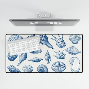 Puede incluir: Alfombrilla de escritorio rectangular con fondo crema e ilustraciones de conchas marinas azules. Un teclado y un ratón blancos están sobre la alfombrilla. La alfombrilla está debajo de un monitor de ordenador.