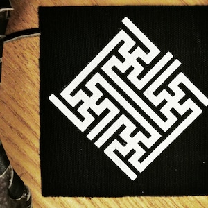 May include: A black fabric patch with a white geometric design resembling a maze or labyrinth. The design is centered on the patch and is a square shape.