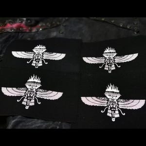 Peut inclure: Quatre patchs en tissu noir avec un motif de créature ailée blanche. La créature porte une couronne et une flamme au-dessus de sa tête. Les patchs sont cousus sur un fond en tissu noir.