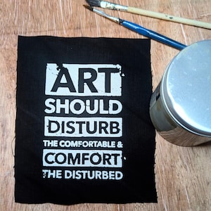 Könnte beinhalten: Schwarzes Stoffquadrat mit dem weißen Text "ART SHOULD DISTURB THE COMFORTABLE & COMFORT THE DISTURBED". Zwei Pinsel und ein silberner Farbtopf befinden sich im Hintergrund auf einer Holzoberfläche.