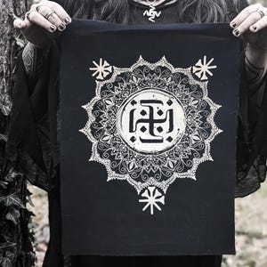 Könnte beinhalten: Ein schwarzes Stoffbanner mit einem weißen Mandala-Design und einem weißen Symbol in der Mitte. Das Symbol ist ein stilisiertes Kreuz mit vier Armen, die jeweils mit einem Kreis enden. Das Mandala ist kompliziert und detailliert, mit einer Vielzahl von Formen und Mustern.