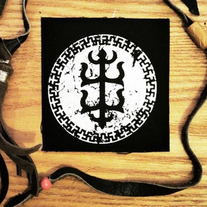 Puede incluir: Parche blanco y negro con un símbolo estilizado desgastado en un círculo. El símbolo es una cruz con un tridente en la parte superior y un corazón estilizado en la parte inferior. El círculo tiene un borde blanco con un patrón repetitivo.