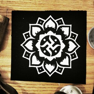 May include: Black fabric patch with a white mandala design. The mandala features a central symbol with four arms and a circle around it. The symbol is surrounded by a ring of petals.