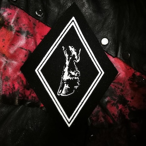 May include: Black and white embroidered patch with a diamond shape and a stylised wolf head inside. The patch is on a black and red background.