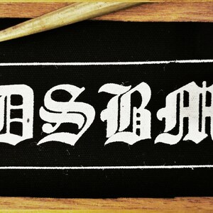 May include: Black fabric patch with white lettering that reads "DSBM" in a gothic font.