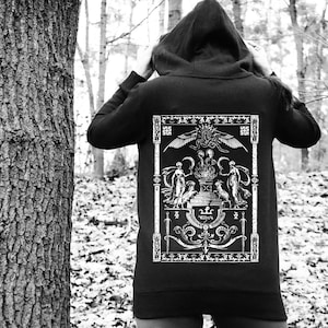 May include: Black hooded sweatshirt with a large white graphic on the back. The graphic features a detailed illustration with figures, wings, and ornate scrollwork. The person is wearing the sweatshirt in a forest setting.