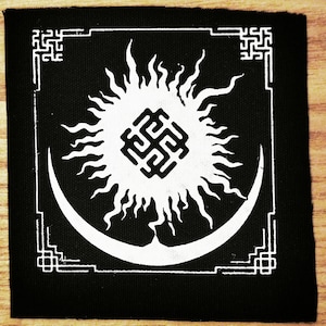 May include: Black and white embroidered patch featuring a sun and moon design. The sun has a stylised sunburst pattern and a square symbol in the centre. The moon is a crescent shape. The design is framed by a square border with decorative corners.