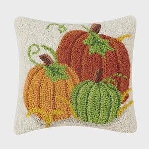 May include: A square throw pillow featuring a textured hooked wool design on an off-white background. The pattern displays three pumpkins in shades of orange, rust, and green, complete with brown stems, green leaves, and curling vines.