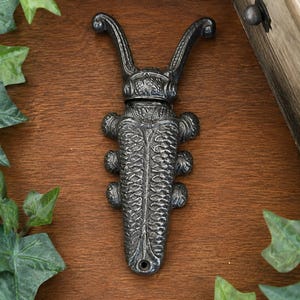 May include: A cast iron boot jack shaped like a beetle with a dark metallic finish. The insect-inspired tool features two long, curved antennae at the top, a textured body with segmented wing covers, and six small legs along the sides. A circular hole is located at the bottom end for mounting or hanging purposes.