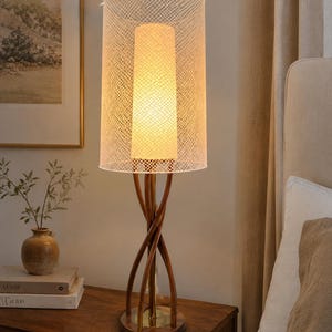 May include: A table lamp featuring a cylindrical white mesh outer shade surrounding a solid inner light diffuser. The base consists of three curved, dark wood supports that twist upward from a circular wooden platform to hold the light fixture.