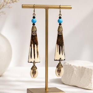 May include: A pair of dangle earrings featuring a triangular bone-like pendant with dark brown vertical streaks, accented by a bright blue bead, a small black bead, and a dangling white cowrie shell, all secured with brass-toned wire wrapping.