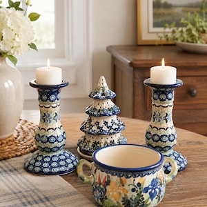 May include: Two ceramic candle holders with a blue and green floral pattern, each holding a white pillar candle. Between them sits a small, perforated ceramic Christmas tree ornament and a two-handled ceramic bowl with a colorful floral design.