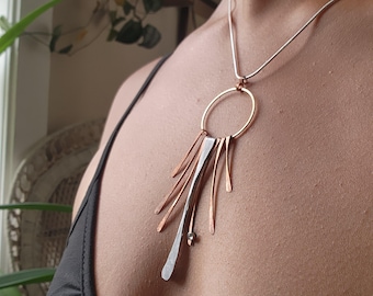 May include: A silver and copper necklace with a circle pendant and a long, thin silver bar with copper accents. The necklace is on a person's chest.
