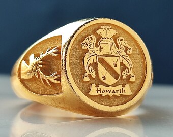 May include: A gold signet ring featuring a circular face engraved with a coat of arms and the name Howarth. The side of the ring displays an engraved stag head. The design includes a shield with a diagonal band and two small stag icons.
