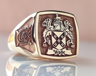May include: A gold signet ring featuring a square face with an engraved coat of arms, including a shield with fleur-de-lis symbols and the name Wade on a banner. The side of the ring displays an engraved letter S with a crown and crossed swords.