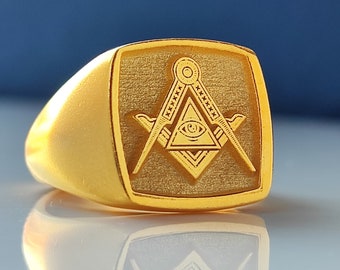 May include: A gold-toned signet ring featuring a square face with a matte finish. The center of the ring displays an engraved Masonic square and compass symbol, which includes an eye within a triangle at its core, creating a detailed emblem.