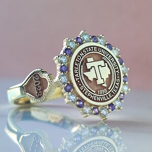 May include: A gold-toned ring featuring a circular emblem for Tarleton State University, Stephenville, Texas, 1899. The emblem is surrounded by alternating purple and clear stones. A heart-shaped detail on the band is engraved with 2023.