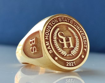 Puede incluir: Un anillo de clase dorado con un sello rojo y dorado de la Universidad Estatal Sam Houston. El sello presenta las iniciales de la universidad "SH" rodeadas de una corona de laurel. El año "2021" está inscrito debajo del sello. El anillo también tiene las letras "BS" grabadas en el lateral.