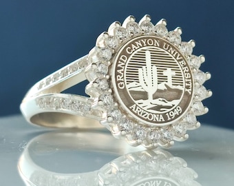May include: A silver ring with a circular design featuring the Grand Canyon University logo and a row of clear stones around the band.