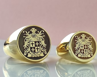 May include: Two gold signet rings featuring a circular face with a dark inlay. Each ring displays an engraved heraldic crest with a shield containing crosses and fleur-de-lis symbols, topped with a helmet, and the name Williams inscribed below.