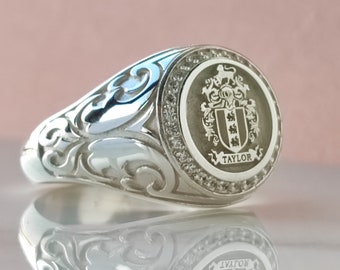 May include: A silver signet ring featuring an intricate scrollwork design on the band and a circular face engraved with a coat of arms and the name TAYLOR. The bezel is surrounded by a textured border, creating a detailed metallic finish.