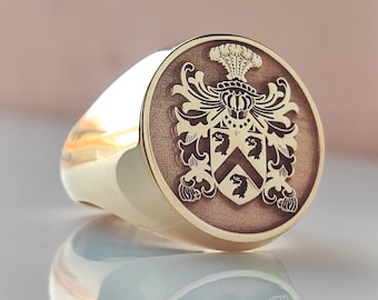 May include: A gold signet ring with a brown crest featuring a shield with a chevron and three birds. The crest is surrounded by ornate floral designs.