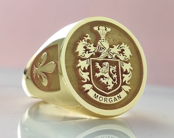 May include: A gold signet ring featuring a circular face with an engraved coat of arms, including a helmet, a lion, and a banner with the text MORGAN. The side of the ring displays a fleur-de-lis emblem. The metal has a polished, reflective finish.