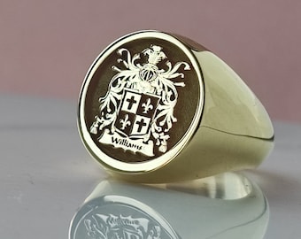 May include: A gold signet ring featuring a circular face with an engraved coat of arms. The crest displays a shield divided into four quadrants with crosses and fleur-de-lis symbols, topped by a helmet, and the name Williams inscribed below.
